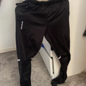 Svarta träningsbyxor från Stanno - Svarta träningsbyxor från Stanno med elastisk midja och dragkedja vid benslut. Byxorna har vita detaljer och är tillverkade i ett syntetmaterial som passar perfekt för sport och träning. Snygg och sportig look med logga på låret.