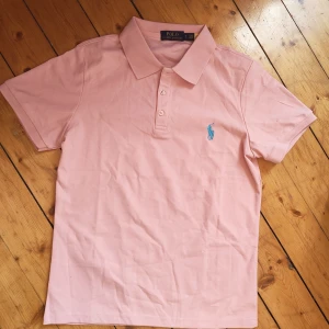 Rosa pikétröja från Polo Ralph Lauren - Snygg ljusrosa pikétröja från Polo Ralph Lauren med klassisk krage och korta ärmar. Tröjan har blå broderad logga på bröstet och är gjord i mjuk bomull. Perfekt för en clean och fräsch look.