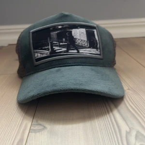 Grön/grå keps från Emilio Pendenza! - Snygg grön/grå keps från Emilio Pendenza, med svartvit fototryck framtill. Kepsen har mesh på sidorna och baksidan för extra ventilation och justerbar snapback. Framsidan är i mjuk manchester och ger en cool vintagekänsla.