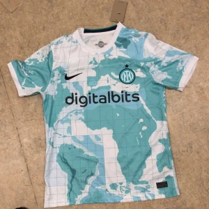 Inter Milan Nike fotbollströja karta - Inter Milan fotbollströja från Nike med unikt kartmönster i vitt och ljusblått. Tröjan har klubbmärke på bröstet, Digitalbits och Lenovo som sponsorer samt svart Nike-logga. Tillverkad i lätt och ventilerande material, perfekt för match eller träning.