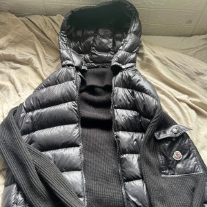 Svart dunjacka från Moncler med huva - Säljer en svart dunväst från Moncler med stickade sidor och ärmar, stor huva och dragkedja framtill. Västen har en snygg ficka med Moncler-logga på ärmen och quiltad design på framsidan. Perfekt för lager-på-lager och streetstyle.