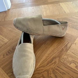 Beige espadrillos från Polo Ralph Lauren - Snygga beige espadrillos från Polo Ralph Lauren i mocka med klassisk broderad logga på sidan. Skorna har rund tå, platt sula och flätad detalj runt sulan. Perfekta för varma dagar och ger en chill vibe till din outfit.