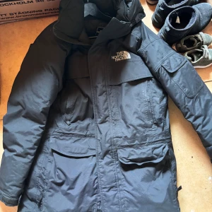 Svart parkajacka från The North Face - Säljer en svart parkajacka från The North Face med flera stora fickor och huva. Jackan är storlek S men passar även M och L. Perfekt för kalla dagar och har en klassisk, stilren design med logga på bröstet. Tillverkad i slitstarkt material och har långa ärmar. Grym för vintern! 