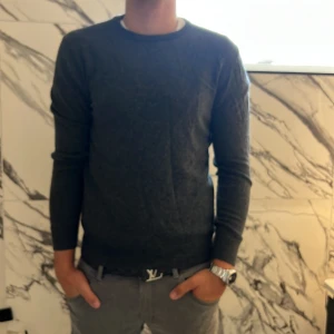 Mörkgrå stickad tröja från Lagency - Säljer en stilren mörkgrå stickad tröja från Lagency i storlek M. Tröjan har rund halsringning, långa ärmar och ribbade muddar vid ärmslut och nederkant. Perfekt för lager-på-lager och enkel att matcha med jeans eller chinos.