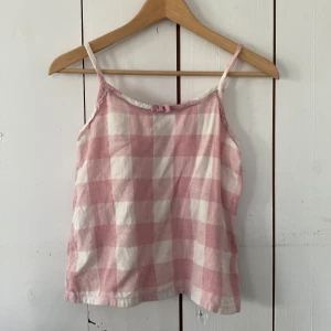 Rutig rosa och vit linnetopp - Supersöt vit och rosa-rutigt pjamassett. Linnet har tunna axelband och en spetsdetalj längst med urringningen med en rosett på. Shortsen har även spetsdetalj, scrunch i midjan och en stor rosett på sig.