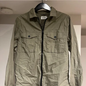 Olivgrön overshirt från East West - Snygg olivgrön overshirt från East West med två bröstfickor med knappar och dragkedja framtill. Långärmad modell i bomull, perfekt att slänga över en t-shirt. Enkel och clean stil som funkar till det mesta.