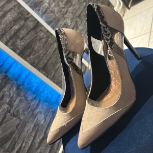 Beige pumps med ormmönster från River Island - Snygga pumps i beige från River Island med spetsig tå och hög klack. Skorna har transparenta detaljer på sidorna och en cool ormmönstrad kant upptill. Materialmix av mocka och syntet ger en modern look. Perfekta för dig som vill sticka ut med stil.