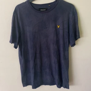 Marinblå t-shirt från Lyle & Scott XXL - Snygg marinblå bommuls t-shirt från Lyle & Scott i storlek XXL. Klassisk passform med korta ärmar och rund hals. T-shirten har ett diskret mönster och den ikoniska gula fågeln broderad på bröstet. Perfekt för en clean och stilren look.