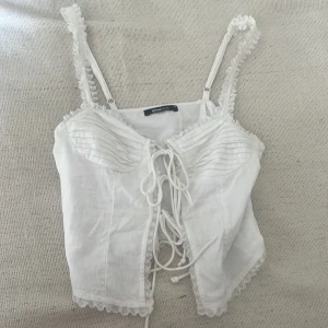Vit korsett-topp från Gina Tricot - Supersöt vit korsett-topp från Gina Tricot med snörning framtill och spetsdetaljer längs kanterna. Axelbanden är smala och dekorerade med volang. Toppen har en romantisk vibe och är perfekt för att lyfta vilken outfit som helst.