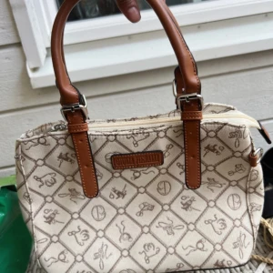 Beige handväska Giulia Pisani mönstrad - Snygg beige handväska från Giulia Pisani med bruna handtag i skinn och coolt rutigt mönster med små symboler. Väskan har dragkedja upptill och en extra ficka med dragkedja på baksidan. Perfekt storlek för det viktigaste och ger en chic vibe.