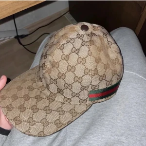 Gucci keps med GG-mönster - Snygg keps från Gucci i beige med klassiskt GG-monogram över hela kepsen. Har en grön och röd rand på sidan som ger den där ikoniska Gucci-looken. Tillverkad i bomullsmix med insida i svart tyg. Perfekt accessoar för att lyfta din outfit.