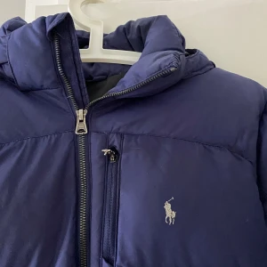 Blå pufferjacka från Polo Ralph Lauren - Snygg blå pufferjacka från Polo Ralph Lauren med broderad logga på bröstet. Jackan har huva, dragkedja framtill och två sidofickor med dragkedja. Perfekt för kalla dagar och riktigt skön att bära. Klassisk och stilren design som funkar till det mesta. strl xl barn (s)