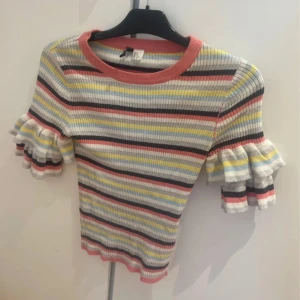 Randig ribbad topp med volangärm H&M strl S - Superfin ribbad topp från H&M med färgglada ränder i rosa, gul, blå, vit och svart. Kortärmad modell med dubbla volanger på ärmarna och rund halsringning. Perfekt för dig som gillar färg och unika detaljer!