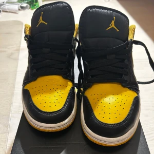 Nike Air Jordan 1 Low svart/gul - Snygga Nike Air Jordan 1 Low sneakers i svart och gul läder med klassisk swoosh på sidan och broderad Jumpman-logga på plösen. Perforerad tåbox för extra ventilation och vit/gul sula som ger en sportig vibe. Perfekta för dig som vill sticka ut med färg. Smått använda.