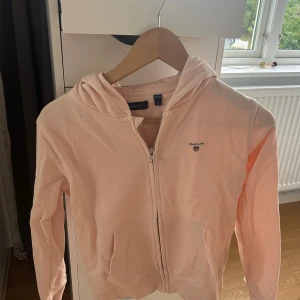 Ljusrosa hoodie från Gant - Säljer en ljusrosa hoodie från Gant med dragkedja framtill och klassisk huva. Tröjan har två fickor fram och Gant-logga broderad på bröstet. Nypris över 700kr.💕Storlek 146/152 men passar XS.