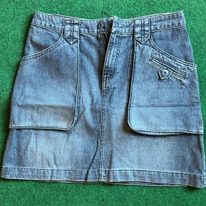 Jeanskjol med stora fickor H&M L.O.G.G. - Säljer en snygg jeanskjol från H&M L.O.G.G. i klassisk blå denim. Kjolen är kort och har två stora fickor framtill, en med knappdetalj. Perfekt för dig som gillar en avslappnad och cool stil.