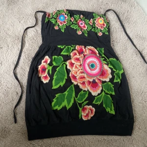 Svart blommig klänning Desigual M - Färgstark kort klänning från Desigual med stora blommor i rosa, rött och grönt på svart botten. Fjäril i grönt på ryggen och knytband upptill. Unik och lekfull design, perfekt för dig som gillar att sticka ut.🌺