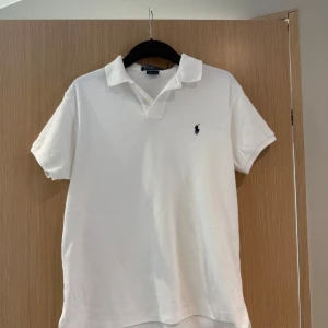 Vit pikétröja från Polo Ralph Lauren - Klassisk vit pikétröja från Polo Ralph Lauren med broderad marinblå logga på bröstet. Tröjan har korta ärmar, krage och knappar framtill. Tillverkad i mjuk bomull som är skön mot huden. Perfekt för en clean och stilren look.