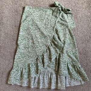 Grön blommig omlottkjol med volang - Supersöt långkjol i grönt med vitt blommönster i storlek 160. Kjolen har omlottmodell med knyt i midjan och volang längs nederkanten.