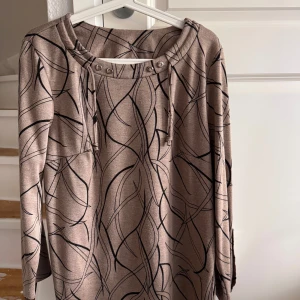 Beige mönstrad långärmad tröja/ tunika  - Säljer en beige långärmad tröja med svart abstrakt mönster. Tröjan har snygga knappdetaljer vid halsringningen och på ärmsluten samt dragsko vid halsen. Perfekt för dig som gillar unika prints . Den motsvarar storlek 46.