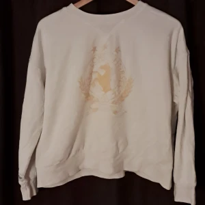 Vit sweatshirt H&M We Love Horses - Vit sweatshirt med tryck i beige och gult framtill, där det står 'We Love Horses' och har en häst i mitten. Tröjan har rund hals, långa ärmar och avslappnad passform. Perfekt för dig som gillar hästar och vill ha en chill look. (Har en patch på höger ärm där det står "Sweden international horse show")