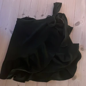 Svart omlottkjol från Vero Moda - Svart kort omlottkjol från Vero Moda med knyt i midjan och volangkant. Bra i passform och gott skick 