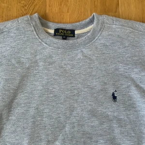Grå sweatshirt från Polo Ralph Lauren - Klassisk grå sweatshirt från Polo Ralph Lauren med broderad marinblå logga på bröstet. Tröjan har rund hals, långa ärmar och ribbade muddar. Tillverkad i mjuk bomullsmix som är skön att bära hela dagen.