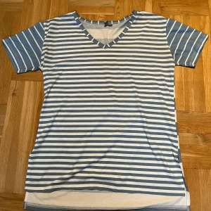 Randig t-shirt från ASOS i blått och vitt - Snygg randig t-shirt från ASOS i blått och vitt med v-ringning och korta ärmar. Skön passform och tillverkad i mjuk bomull, perfekt för en chill och stilren look. Klassiska smala ränder framtill och bredare ränder baktill ger en unik twist.
