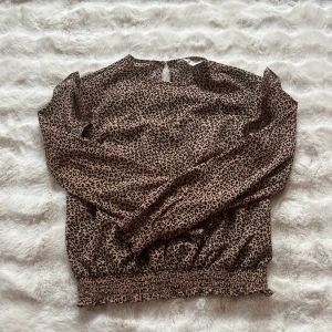 Leopardmönstrad blus med volangärm - Snygg blus med leopardmönster i beige och svart. Långärmad modell med volangdetaljer på axlarna och smock vid ärmslut och midja. Knäppning bak i nacken med liten knapp. Tunt, flowy material som känns luftigt och trendigt.