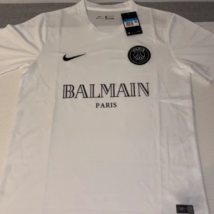 Balmain x PSG T-shirt – vit & helt ny - Obs! Aldrig använd, taggad med originaletikett. Stilren träningstisha med både Paris Saint‑Germain‑logo och “Balmain” på bröstet.