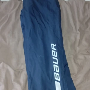 Blå täckbyxor från Bauer, M - Mörkblå täckbyxor från Bauer med resår i midjan och grått innerfoder. Byxorna har en sportig look med Bauer-loggan tryckt längs benet. Perfekta för vinterns aktiviteter och håller dig varm och torr.