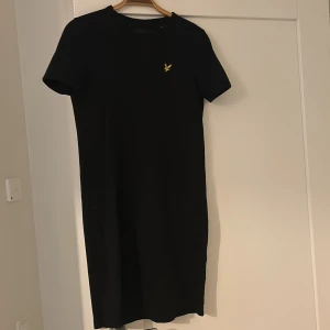 Svart t-shirtklänning Lyle & Scott XS - Svart t-shirtklänning från Lyle & Scott i 100% ekologisk bomull. Enkel och stilren design med kort ärm och rund hals. Liten gul logga på bröstet ger en snygg detalj. Perfekt för en avslappnad och cool look.