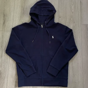 Marinblå hoodie från Polo Ralph Lauren - Snygg marinblå hoodie från Polo Ralph Lauren med dragkedja och huva. Klassisk logga broderad på bröstet . Storlek M, hör av er vid frågor med mera.
