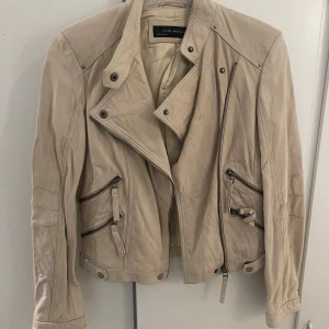 Beige skinnjacka från Zara Woman - Snygg beige skinnjacka från Zara Woman med coola detaljer som snedställda dragkedjor och tryckknappar. Jackan har lång ärm, klassisk bikerkrage och flera fickor framtill. Perfekt för dig som vill ha en stilren men edgy look.