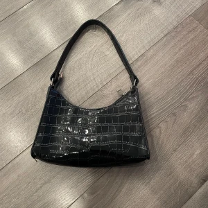 Svart axelväska i croco-imitation - Snygg svart axelväska med glansig croco-imitation och stilren siluett. Väskan har ett kort axelband och stängs med dragkedja upptill. Perfekt accessoar för att lyfta din outfit.