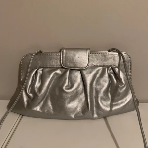 Silver axelväska från Accessories by Wedins - Snygg axelväska i metallic silver från Accessories by Wedins. Väskan har ett veckat framparti, lång axelrem och stängs med ett lock. Perfekt för dig som vill sticka ut med en glansig accessoar. Rymmer mobil, plånbok och småsaker. Väskan har några små defekter och märken så skriv om frågor!