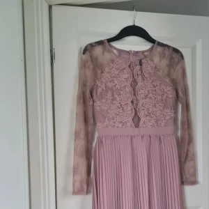 Ljusrosa långklänning med spets och plisserad kjol - Säljer en elegant ljusrosa långklänning med spetsdetaljer upptill och plisserad kjol. Klänningen har långärmade spetsärmar och en rund halsringning. Perfekt för speciella tillfällen..Om det behövs kan jag skicka bättre bilder på klänningen vid intresse 