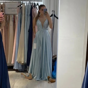 Ljusblå långklänning med djup v-ringning - Säljer en elegant ljusblå långklänning med djup v-ringning och öppen rygg. Klänningen har smala axelband och ett fint fall i kjolen. Perfekt för bal eller festliga tillfällen. Materialet är glansigt och känns lyxigt. Jag är 163 cm och har klackar på bilderna. Klänningen är lätt uppsydd och går enkelt att lägga ned igen. Bara använd en gång. Storlek 0