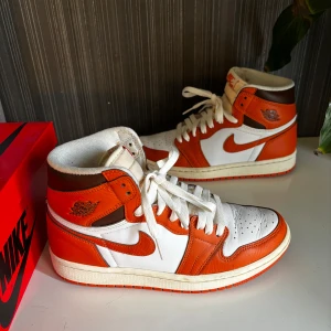 Nike Air Jordan 1 High OG orange och vita - Säljer ett par Nike Air Jordan 1 High OG i orange, vitt och brunt skinn. Klassisk high-top siluett med vit bas, orange overlays och bruna detaljer. Tjock vit sula och orange yttersula. Perfekta för dig som gillar sneakers med retrovibe.