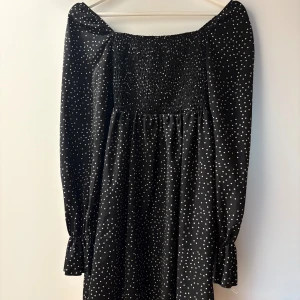 Svart prickig klänning med smock och långa ärmar - Svart klänning med vita prickar, smockad upptill och långa ärmar med volang vid ärmslut. Klänningen har en rak passform och är 100% polyester. Kort klänning 👗 