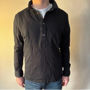 J.Lindeberg piké - Säljer en svart långärmad piké med klassisk krage och knäppning framtill. Skjortan har en avslappnad passform och är perfekt att styla med jeans eller chinos. Enkel och stilren design som funkar till många tillfällen.