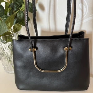 Svart väska - Svart väska med gulddetaljer och dubbla handtag. Väskan har flera fack inklusive ett med dragkedja, och stängs med magnetknappar. 