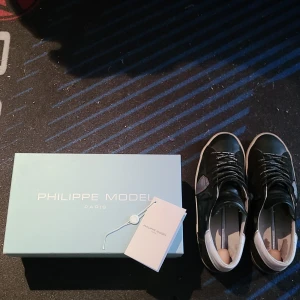 Philippe Models - Säljer ett par svarta Philippe Model sneakers med vita och grå detaljer. Skorna har snörning, rund tå och platt sula. Märkeslogga syns på sidan och bak på hälen. Perfekta för dig som gillar stilrena och exklusiva sneakers. Dom är lite slitna i hälen under som man ser kan skicka bättre bild men man känner inte det alls det läcker ej in nå vatten osv... riktigt fina skor enligt mig fortfarande bra skick. Har lagat skadorna själv med gorilla glue och sånt, har fungerat utmärkt. 