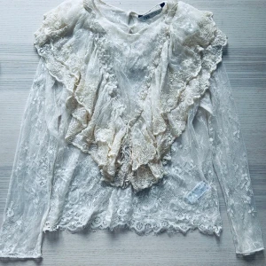 Vit spetsblus med volang från Zara - Romantisk vit spetsblus från Zara med långa ärmar och vackra volangdetaljer framtill och vid axlarna. Blusen är genombruten och har ett feminint uttryck med mycket spets och fina detaljer.