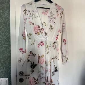 Vit blommig långklänning med v-ringning - Säljer en romantisk vit långklänning med blommigt mönster i rosa, lila och grönt. Klänningen har v-ringning, långa ärmar och markerad midja. Perfekt för sommarens alla tillfällen!