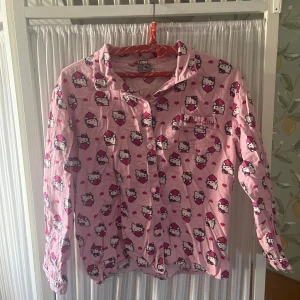Rosa Hello Kitty skjorta - Säljer en söt rosa skjorta med Hello Kitty-motiv över hela plagget. Skjortan har långa ärmar, klassisk krage och knäppning framtill. Perfekt för dig som älskar gulliga och lekfulla plagg! Officiell Sanrio merch!!