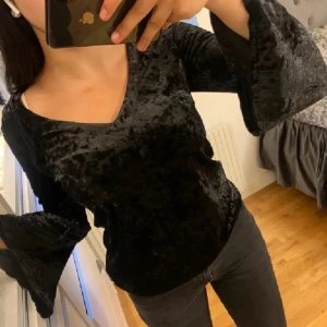 Svart sammetsblus med utsvängda ärmar - Säljer en svart sammetsblus med v-ringning och vida, utsvängda ärmar. Blusen har en snygg knytning i ryggen och är perfekt för dig som vill ha något lite extra. Materialet är mjukt och glansigt. Från Capri Collection storlek Xs.