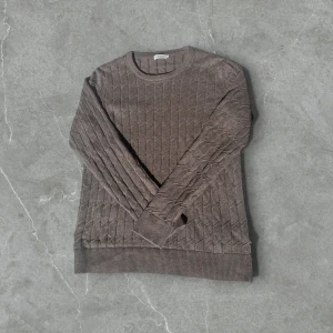 Brun stickad tröja från J.Lindeberg - Säljer en brun stickad tröja från J.Lindeberg med diskret rutmönster. Tröjan har rund halsringning och långa ärmar, perfekt för kyligare dagar. Klassisk och stilren design som passar till mycket.