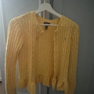  kabelstickad kofta från Polo Ralph Lauren - Säljer en söt gul kabelstickad kofta från Polo Ralph Lauren i storlek XL/16, men passar som en S. Inte använd mycket och därför mycket fint skick🎀Den har väldigt fina volangdetaljer nedtill. Jättefin till sommaren eller med ett par ljusa byxor💗