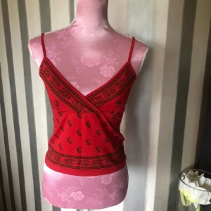 Röd fejkomlott linnetopp med mönster - Säljer en röd omlott linnetopp med tunna axelband och svart blommigt mönster. Toppen har en dekorativ bård längs nederkanten och omlottdesign framtill. Perfekt för varma dagar och enkel att styla.